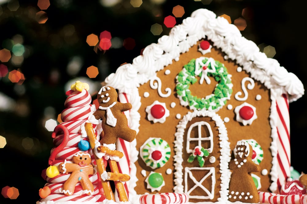 A gingerbread house with Christmas lights in the background (very shallow DOF with focus on the gingerbread man decorating the candy cane tree in front of the gingerbread house).