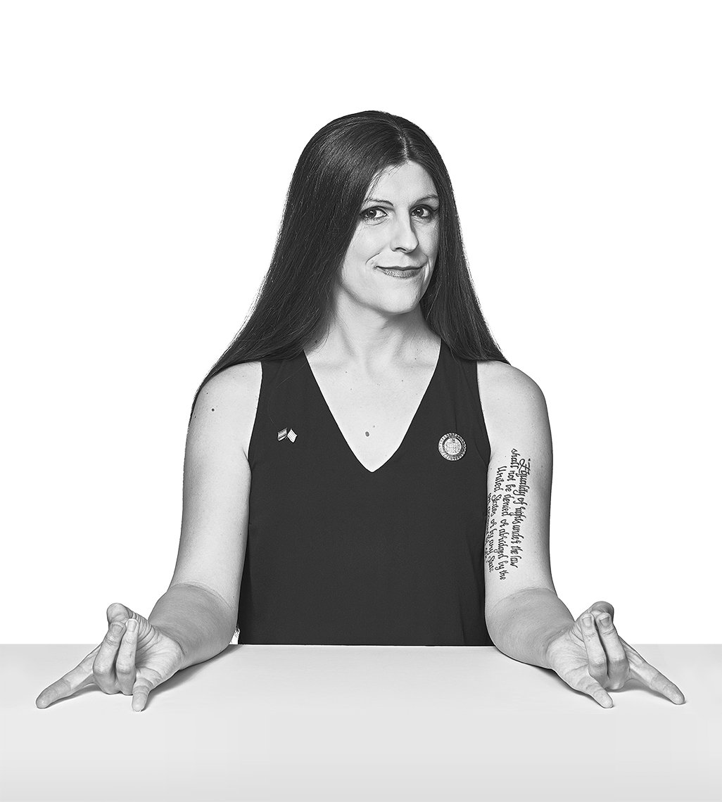 Portrait of Danica Roem with her arms leaning against a table, both hands making the rock-on-hand gesture.