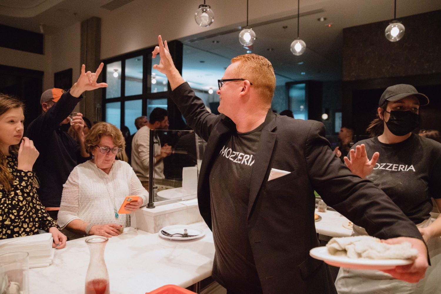 A man with ginger hair and rectangle glasses in a black shirt and blazer says "I love you" in American Sign Language to a customer at a restaurant, who signs "I love you" back.