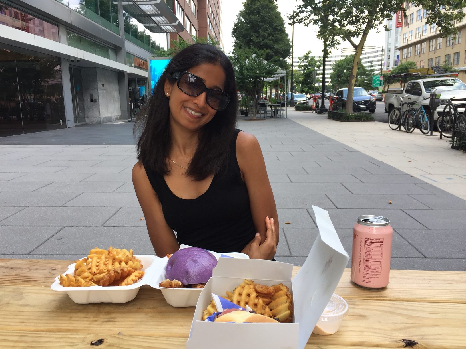 A woman with black hair wearing large dark glasses smiles at the camera. A purple burger, waffle fries, and a pink can sit on the table in front of her.