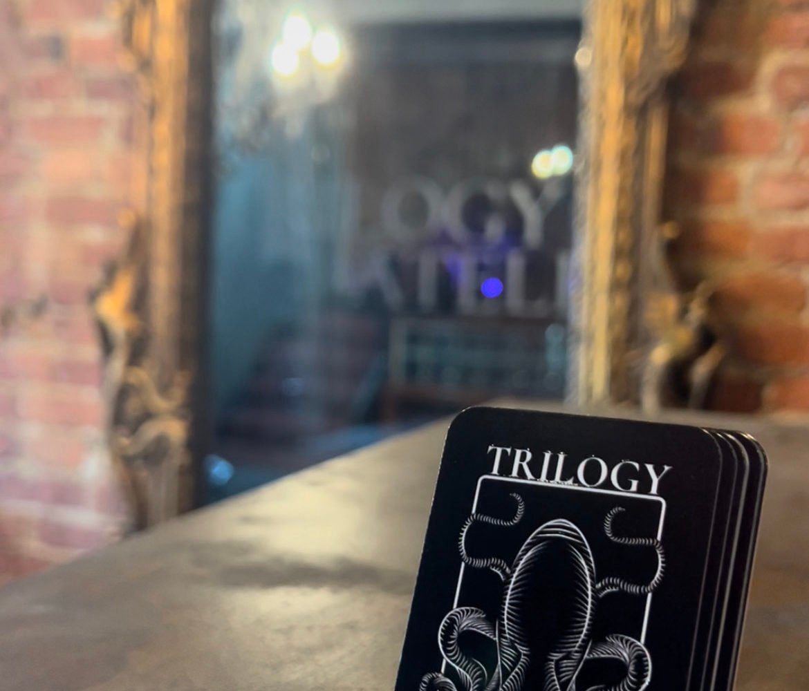 Black and white business cards sit in a holder on a table. The business cards say "Trilogy Atelier" and have a kraken logo in minimalist black and white. An ornate gold mirror is behind the business cards, and reflects the glass doors which have "Trilogy Atelier" printed on them in white.