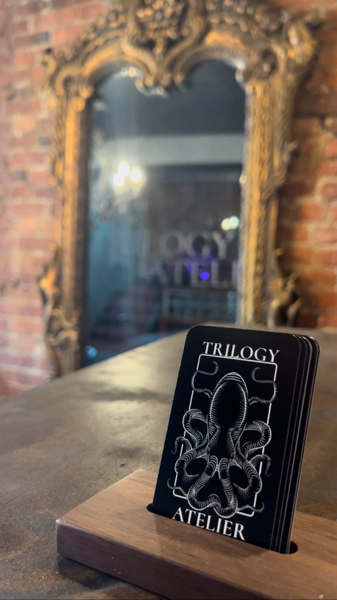 Black and white business cards sit in a holder on a table. The business cards say "Trilogy Atelier" and have a kraken logo in minimalist black and white. An ornate gold mirror is behind the business cards, and reflects the glass doors which have "Trilogy Atelier" printed on them in white.