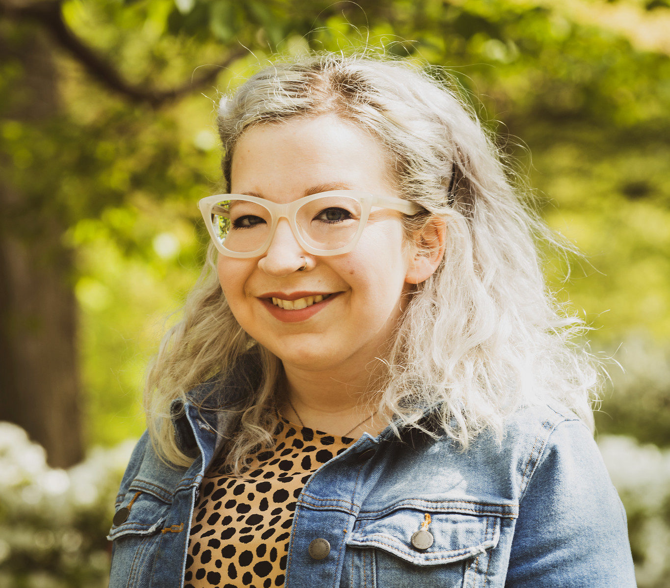 A woman with platinum blond hair, white-framed glasses wearing a leopard print shirt under a blue denim jacket smiles at the camera. 