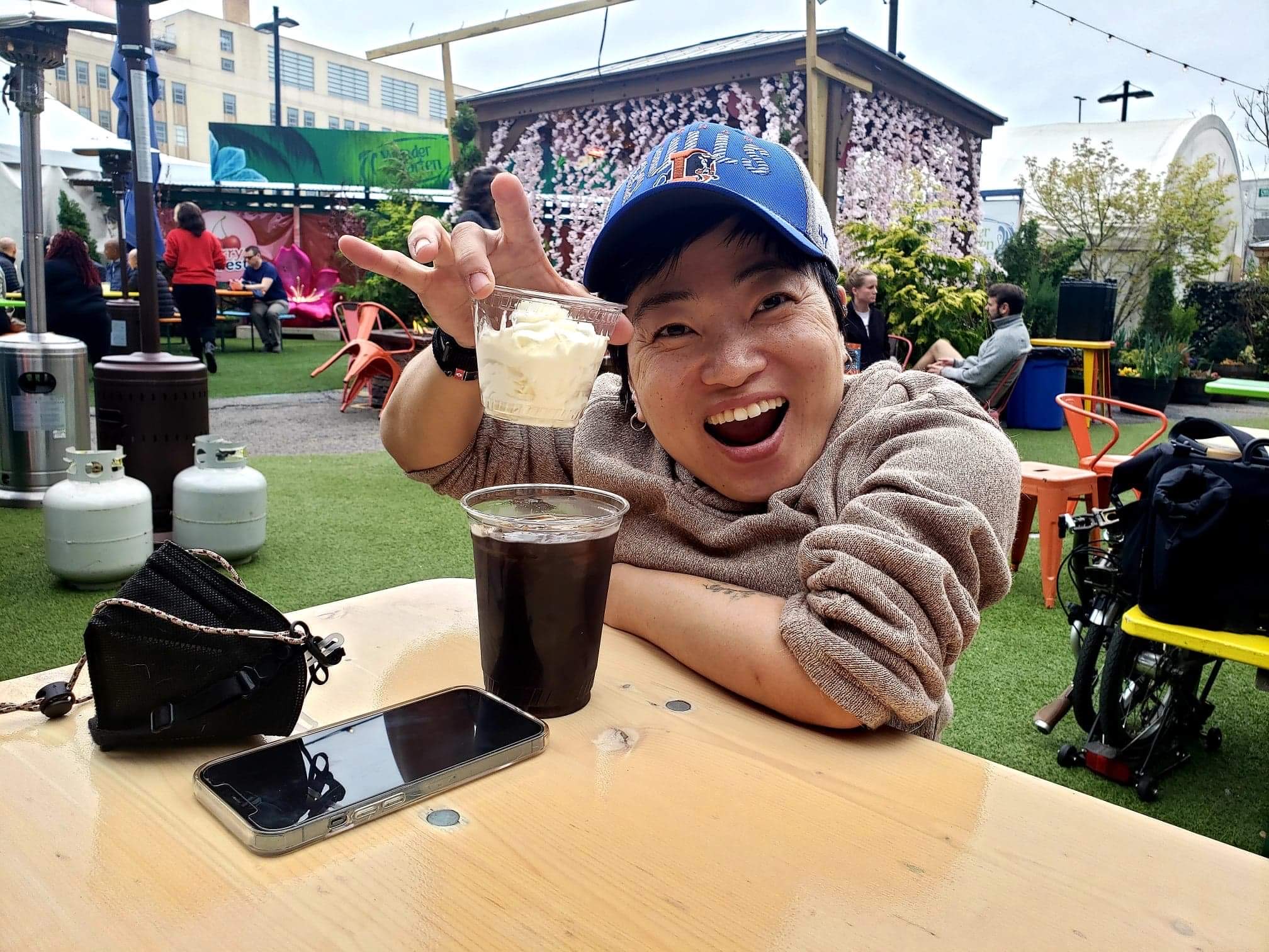 A woman in a baseball cap leans against a table, smiles at the camera and holds up a drink.