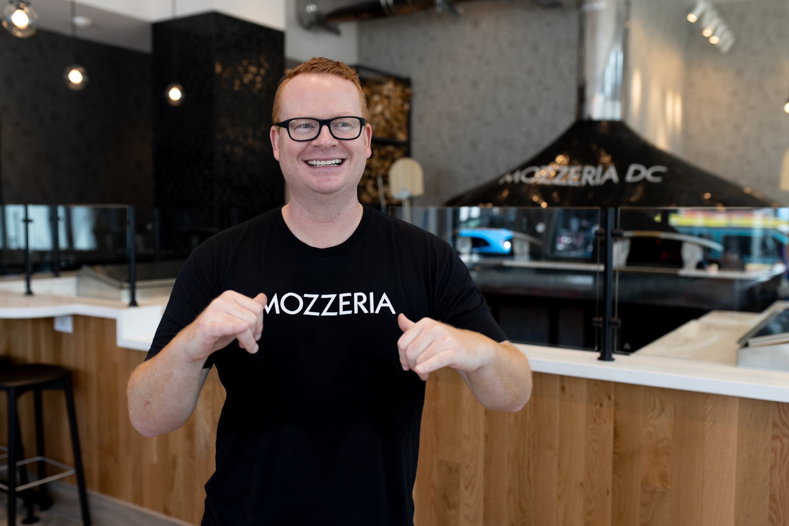 A man with ginger hair and rectangular glasses smiles at the camera. He is wearing a black shirt that says "Mozzeria" on it.