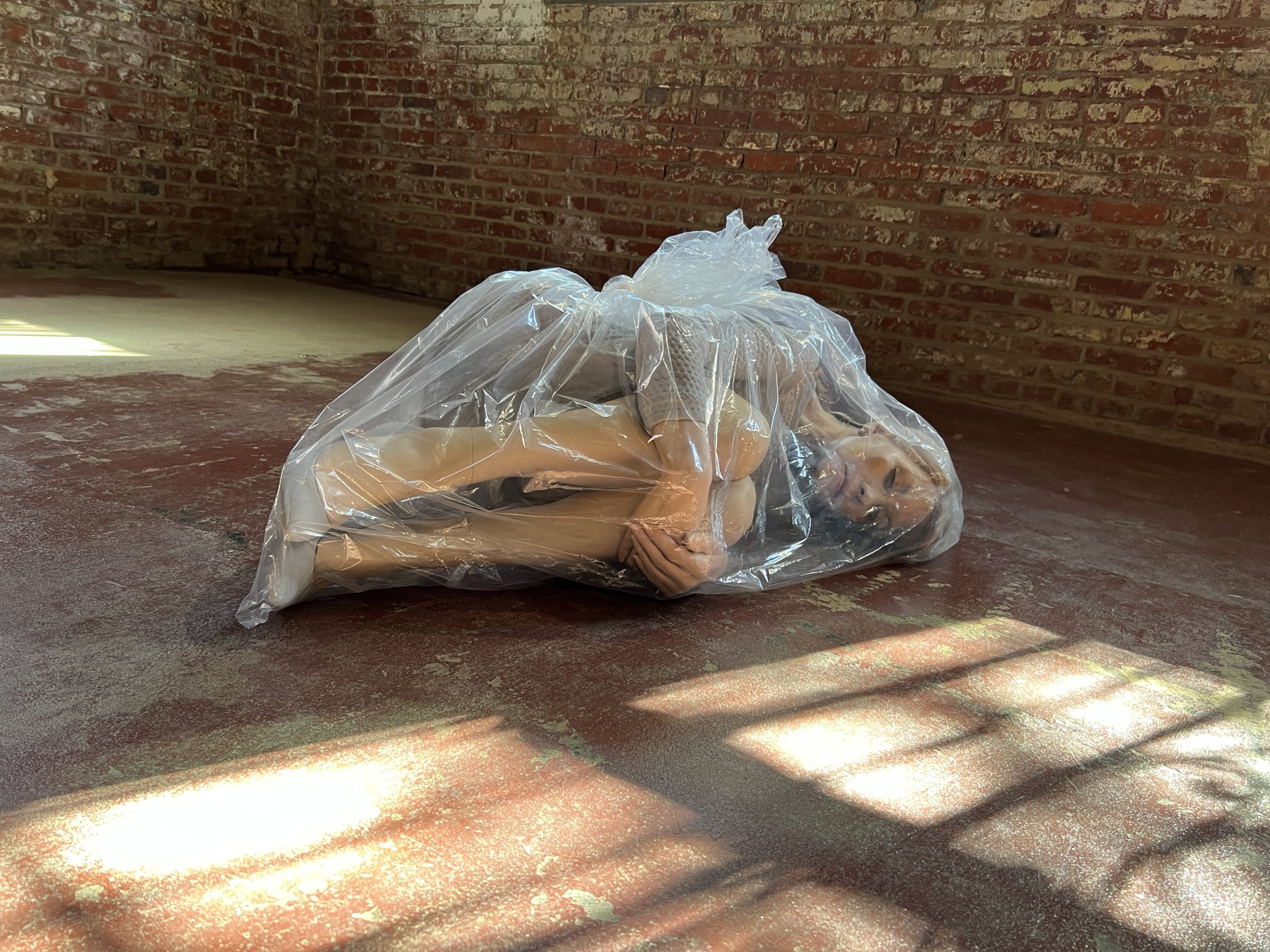 A plastic doll lays in the fetal position inside of a plastic garbage bag