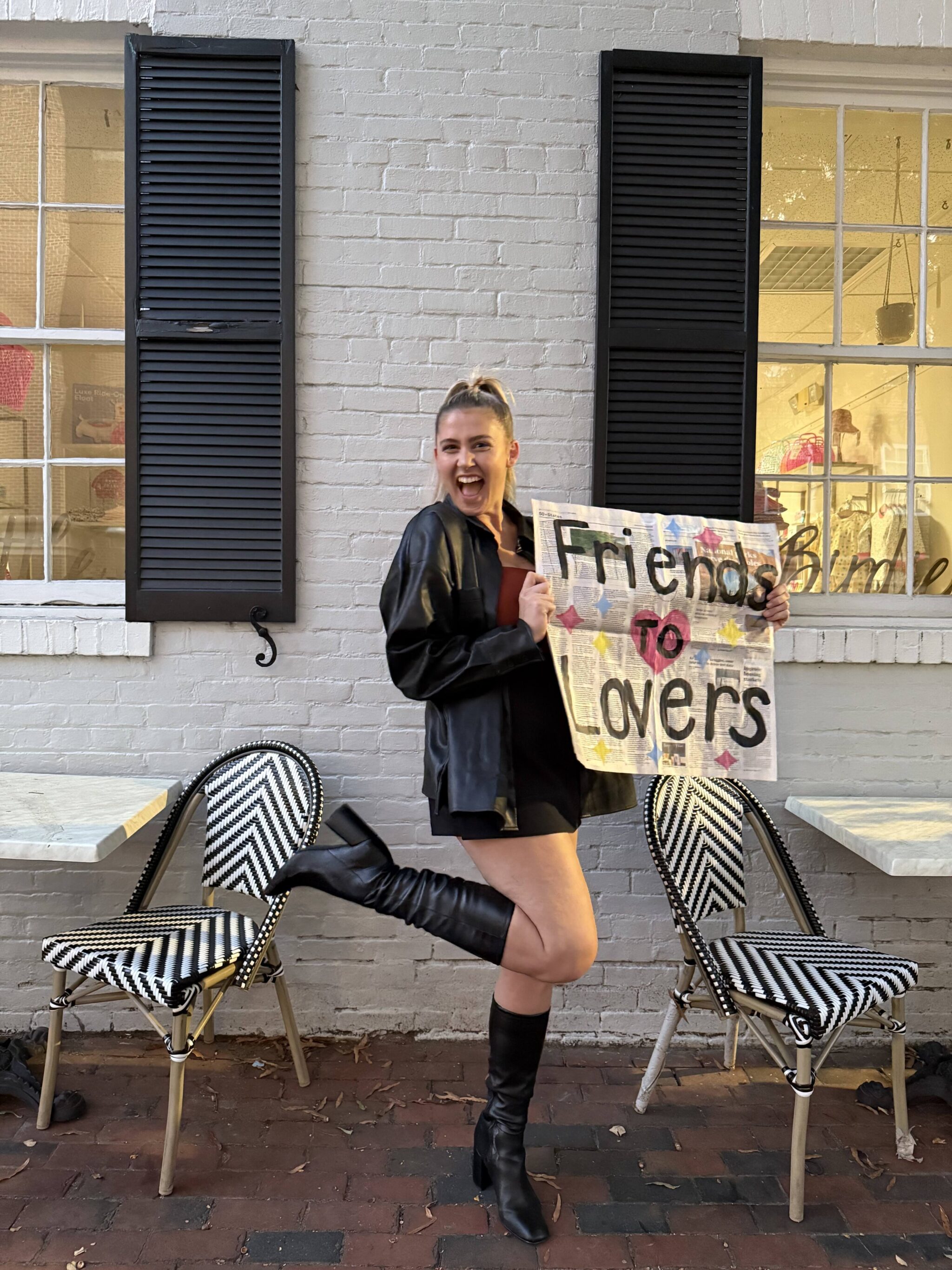 A blonde woman wearing a black coat and boots smiles and holds up a sign that says "Friends to Lovers."
